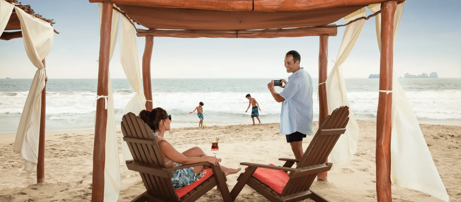 woman sitting in cabana chair while man takes photo of children playing in sand