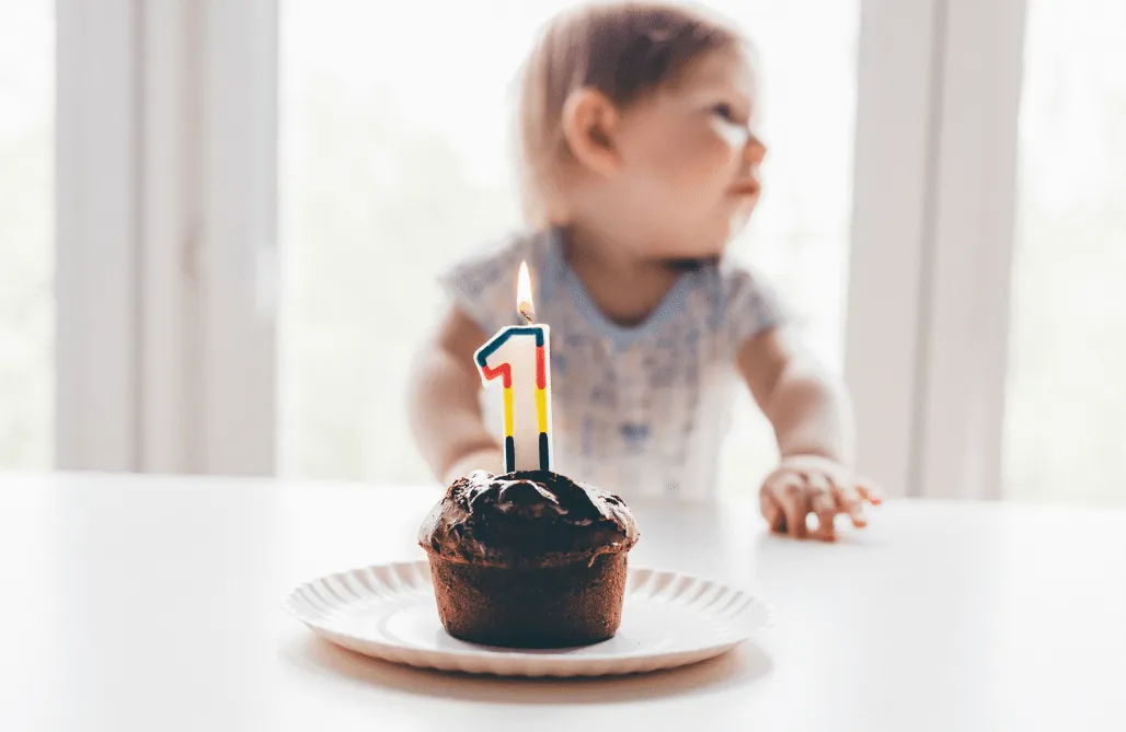 Chocolate cupcake with lit number one candle and baby in background.
