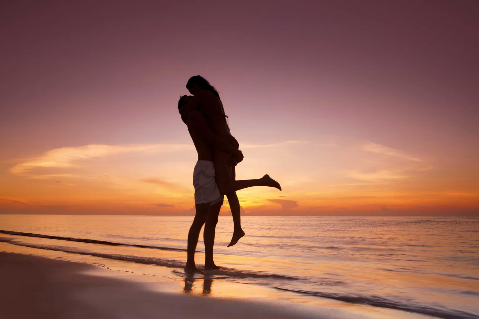man picking up significant other and embracing on shoreline during sunset