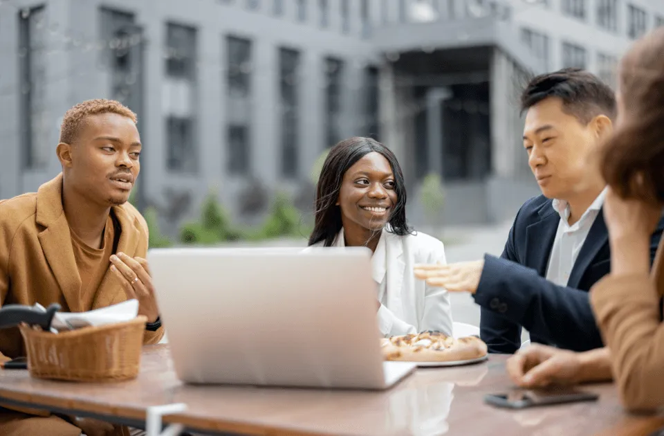 A group of coworkers sitting outdoors at a table, smiling and talking while looking at a laptop, with food and drinks nearby.