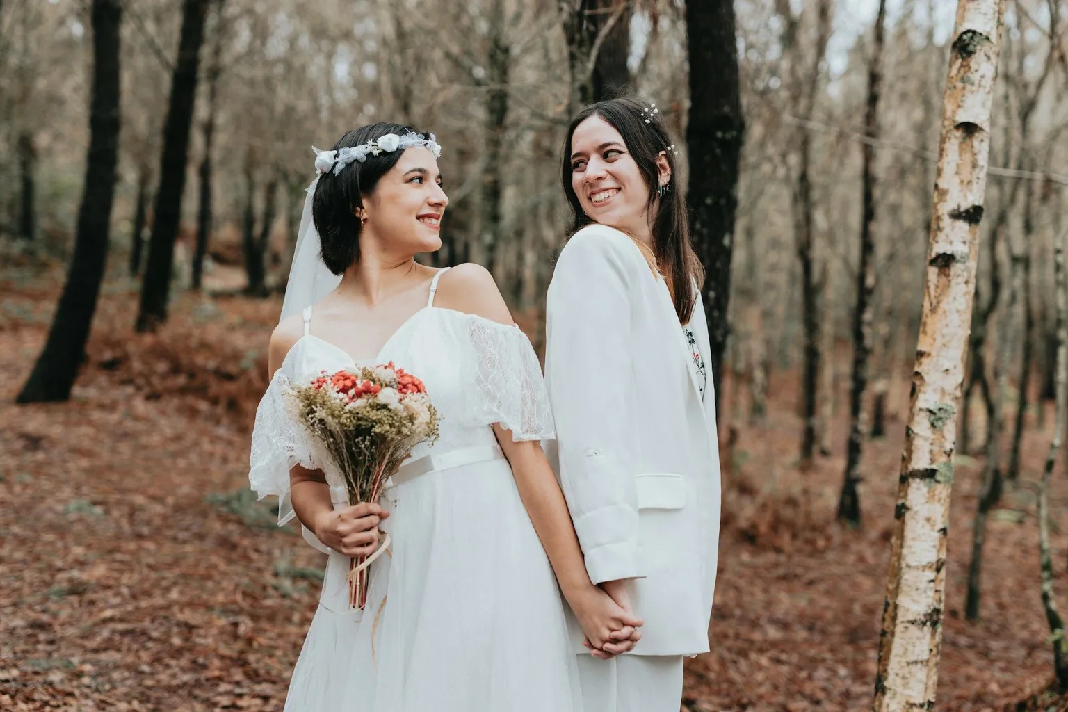 Two brides holding hands and smiling at each other in a forest wedding.