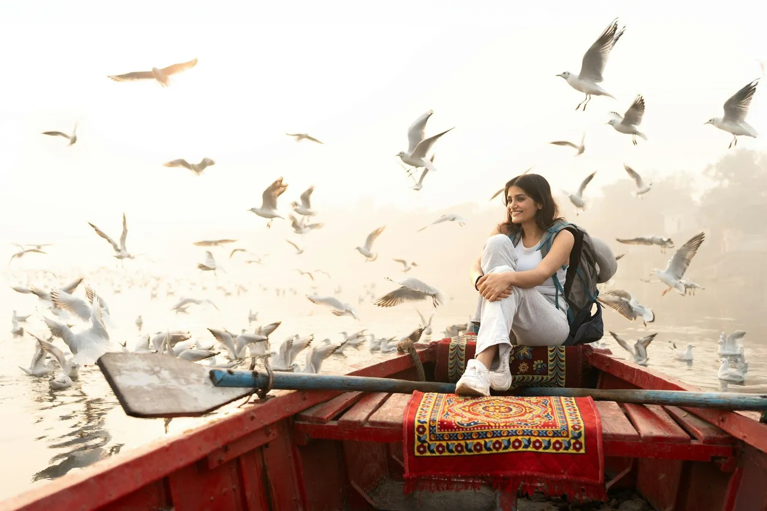 Woman sitting on a red boat surrounded by seagulls at sunrise.