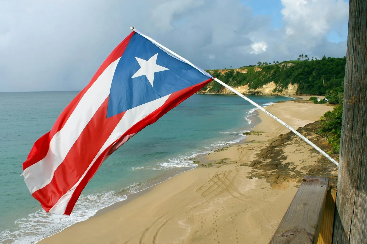 Puerto Rico flag flying above a sandy beach and turquoise ocean along a tropical coastline.