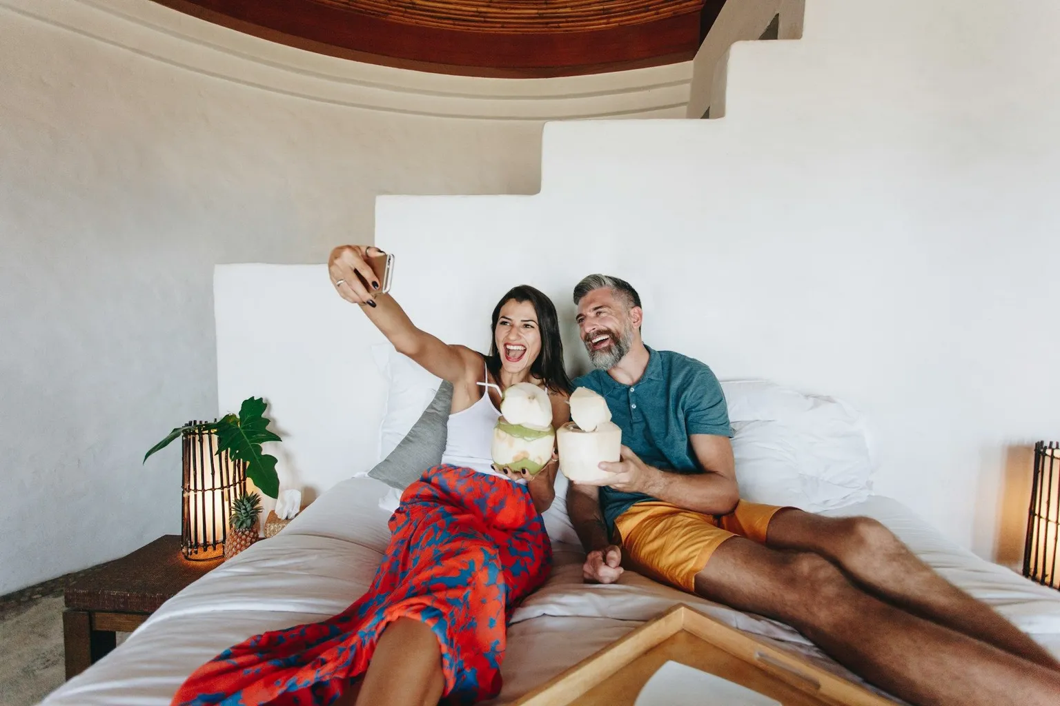 Smiling couple taking a selfie with fresh coconuts on the bed of a bright white honeymoon villa.