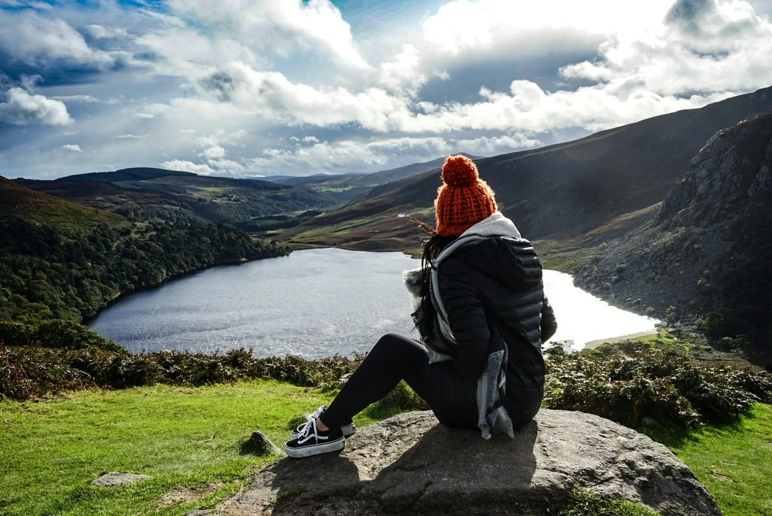 Hiker in an orange beanie sitting on a rock overlooking a lake and green valley in the Irish countryside.