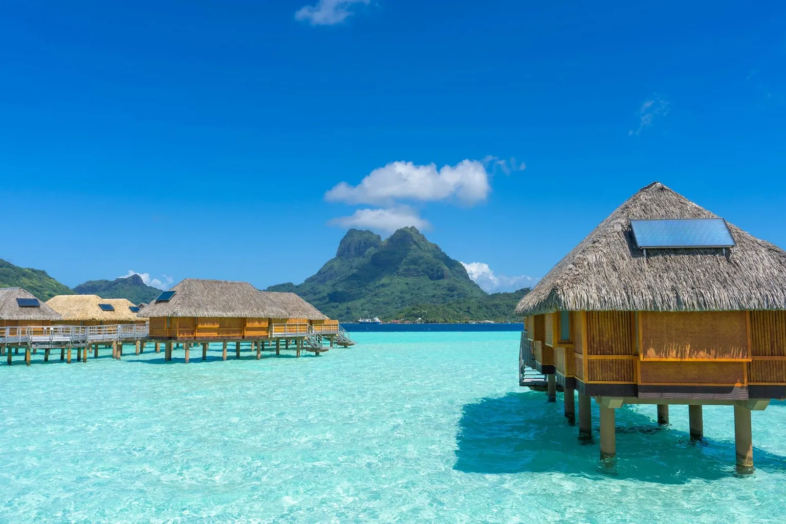Overwater bungalows in Bora Bora with turquoise lagoon and a mountain under a clear blue sky.