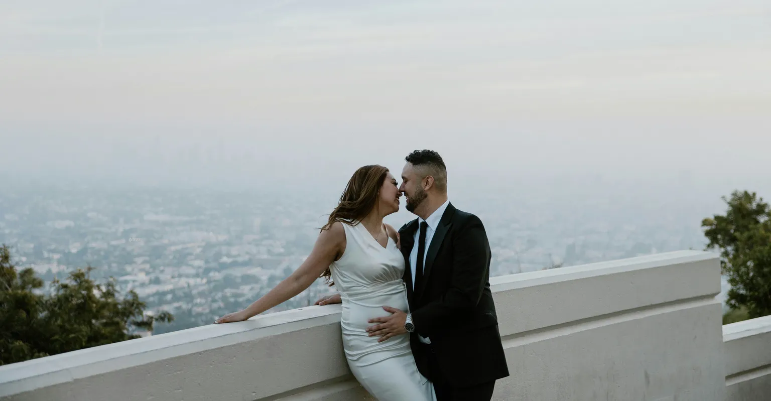A bride and groom share a kiss while overlooking a hazy city skyline at sunset.