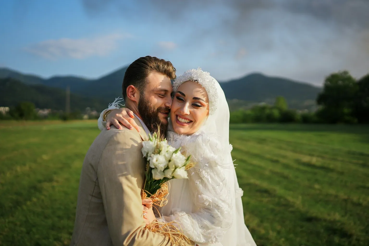 Smiling bride and groom hugging in a green field with mountains in the background.