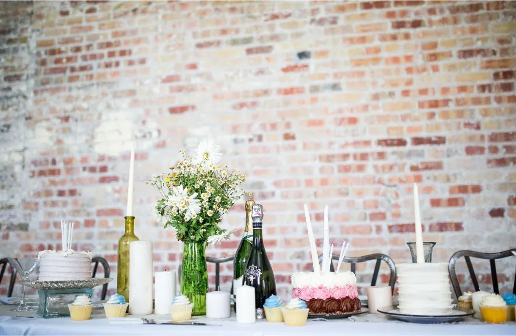 Decorated dessert table with cakes, champagne, candles, and flowers against a brick wall.