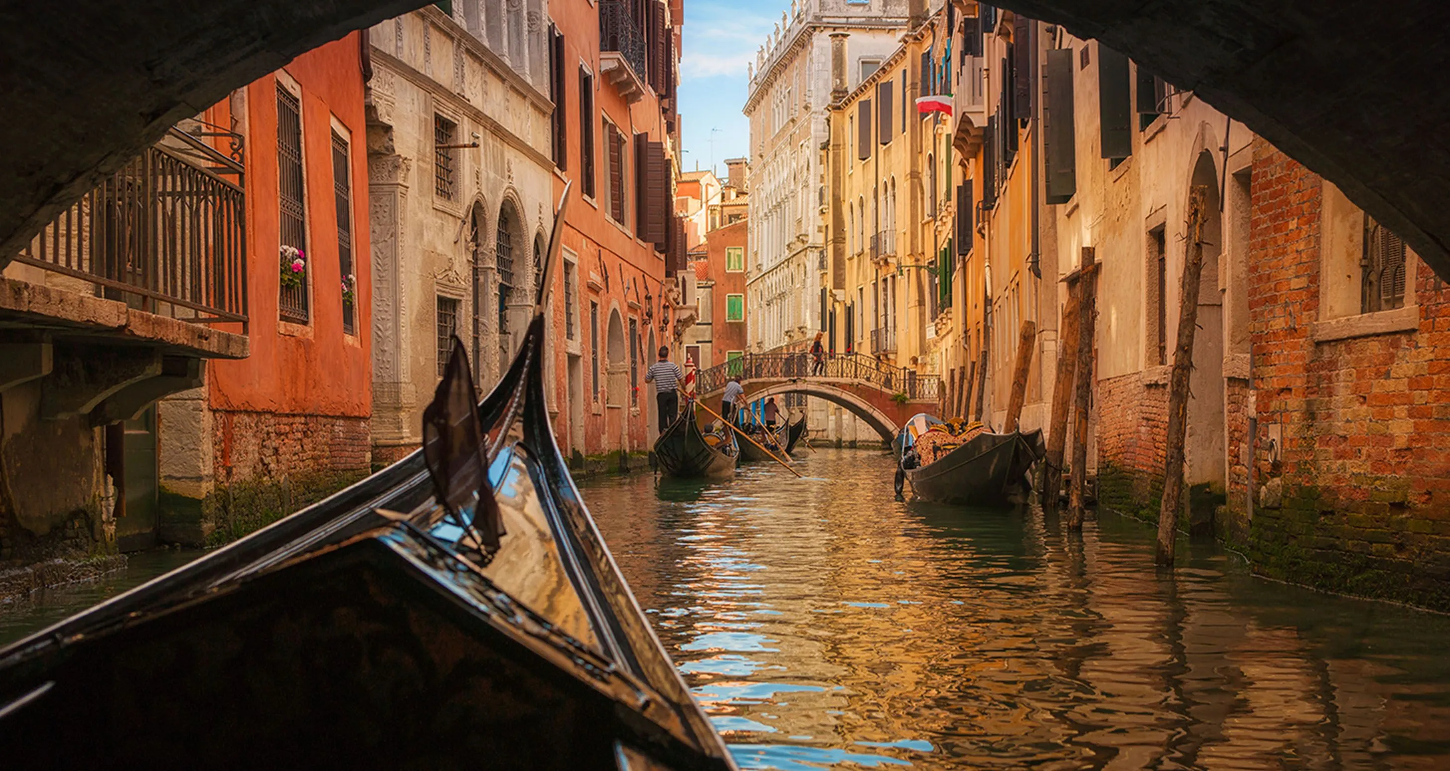 Gondolas moving through a narrow canal in Venice with historic buildings and a small bridge in the background.