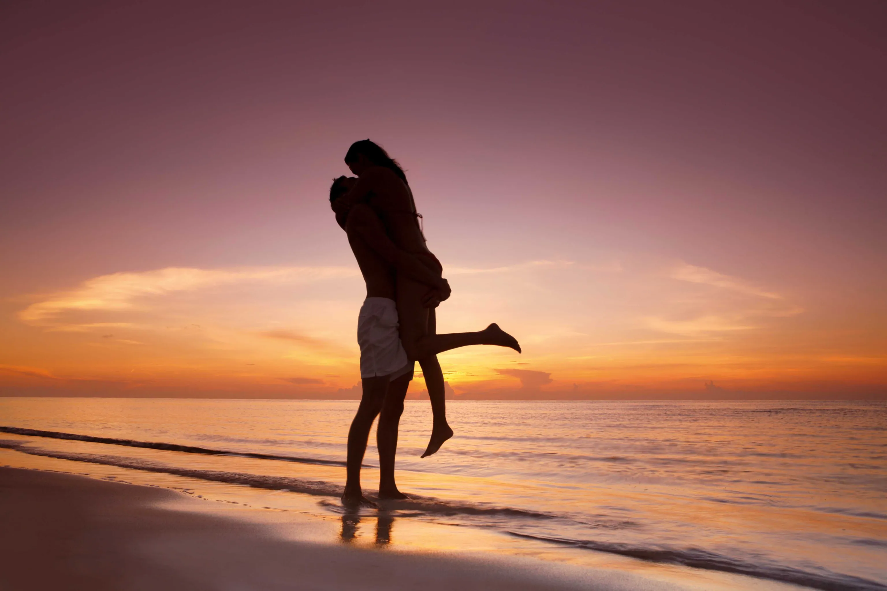 man picking up significant other and embracing on shoreline during sunset