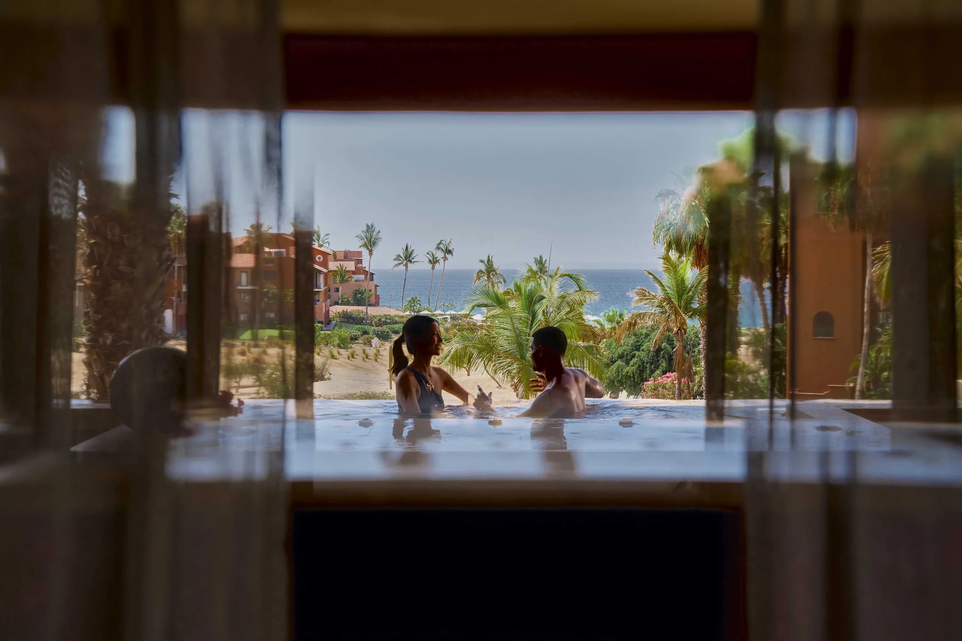 A couple relaxing in an infinity pool overlooking a tropical ocean with palm trees and a sandy beach