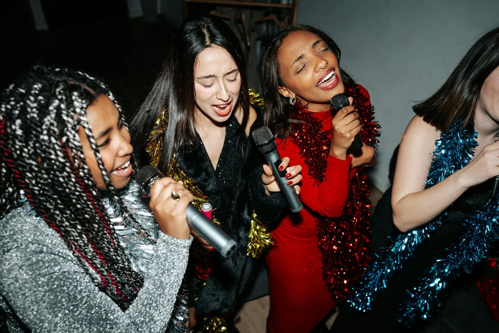 Four women singing karaoke together while holding microphones at a party.