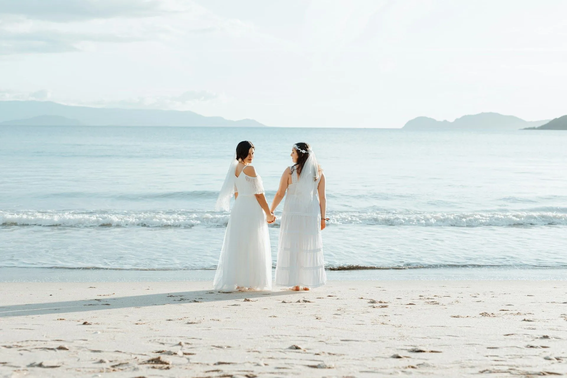 two brides holding hands on shoreline with ocean in background