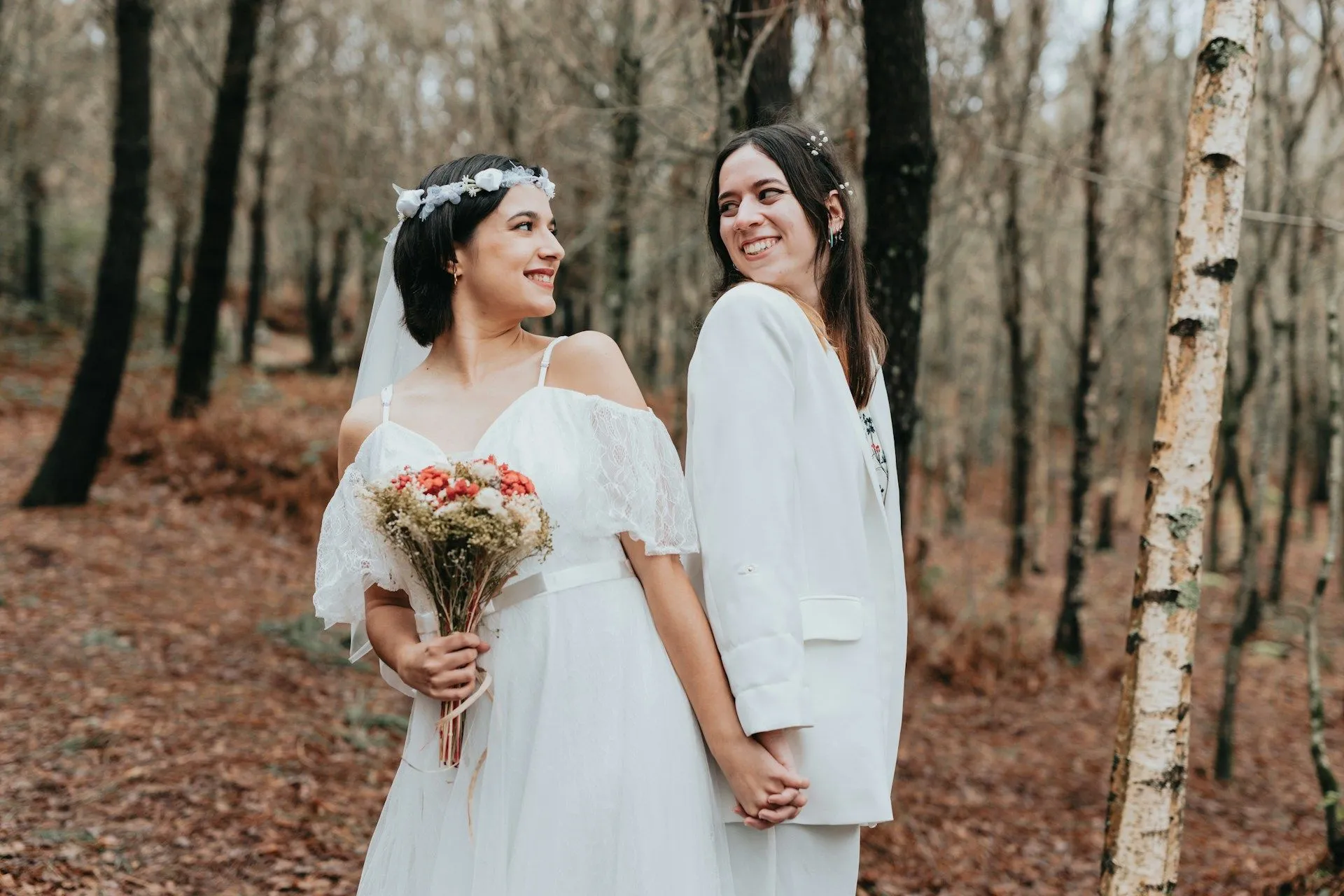 Two brides holding hands and smiling at each other in a forest wedding.