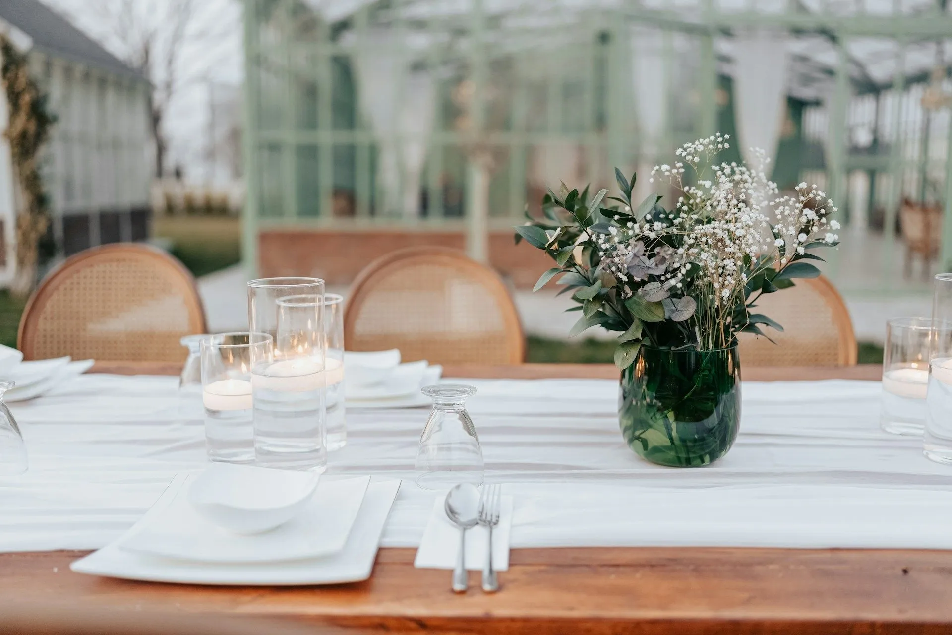 Elegant outdoor table setting with white linens, candles, and greenery.