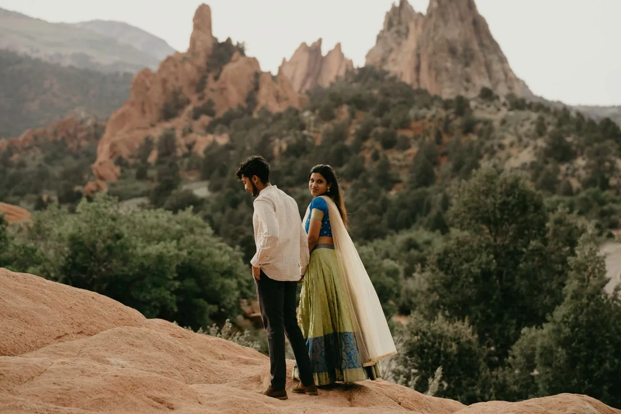 Couple standing on a rocky overlook with red rock formations and greenery in the background.