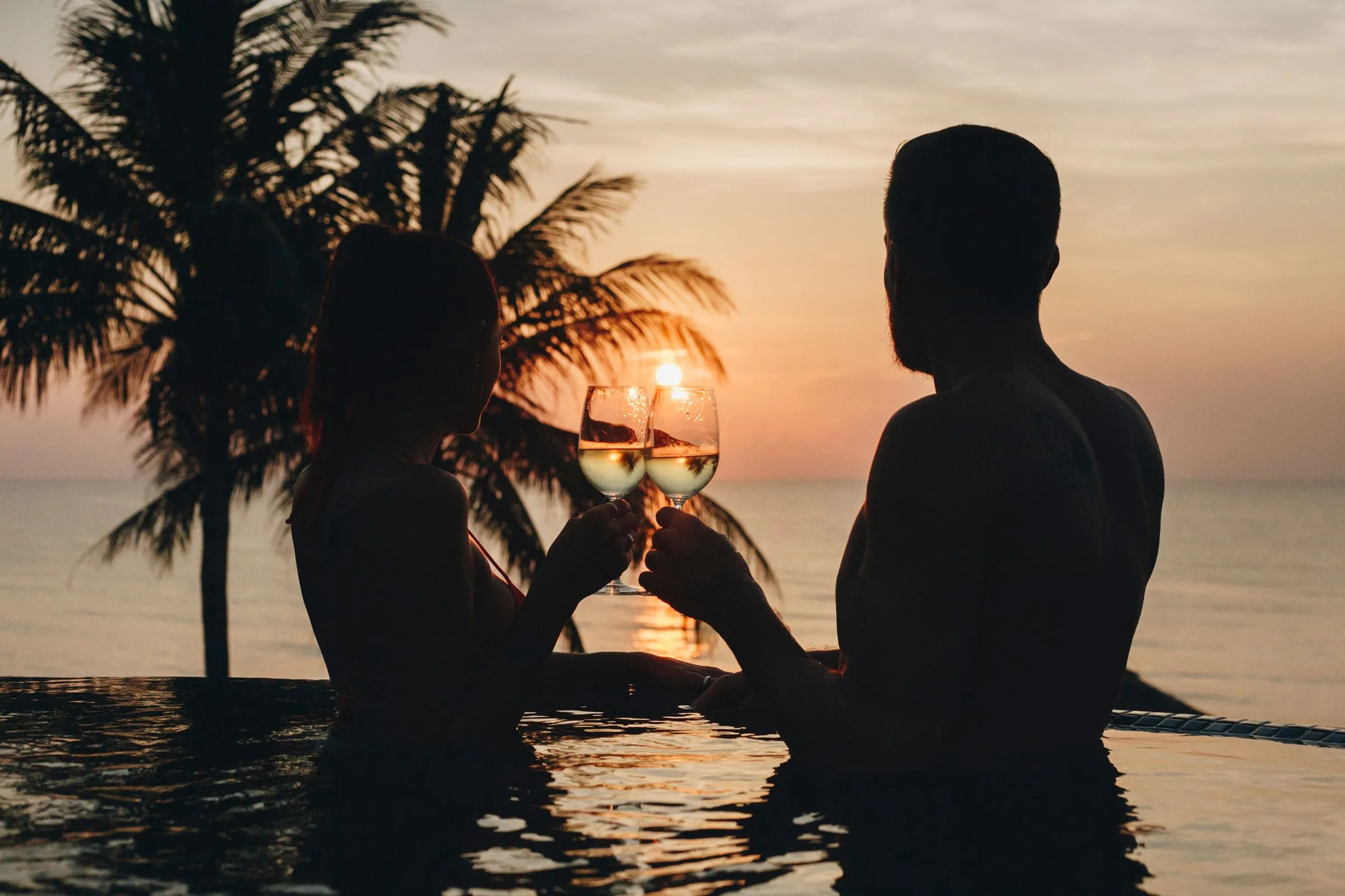Couple clinking glasses in a pool at sunset, with palm trees and the ocean in the background.