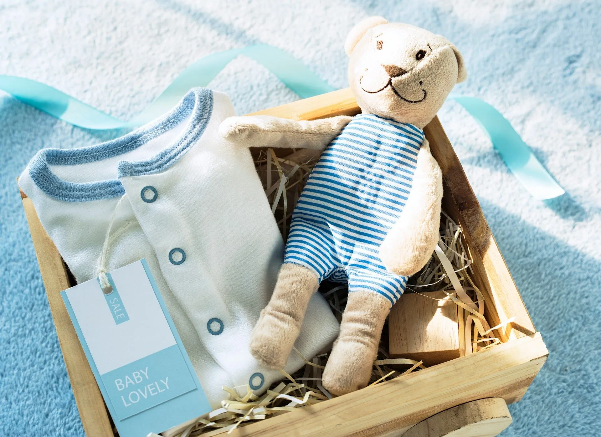 A baby sprinkle gift box with a onesie and striped teddy bear on a blue blanket.
