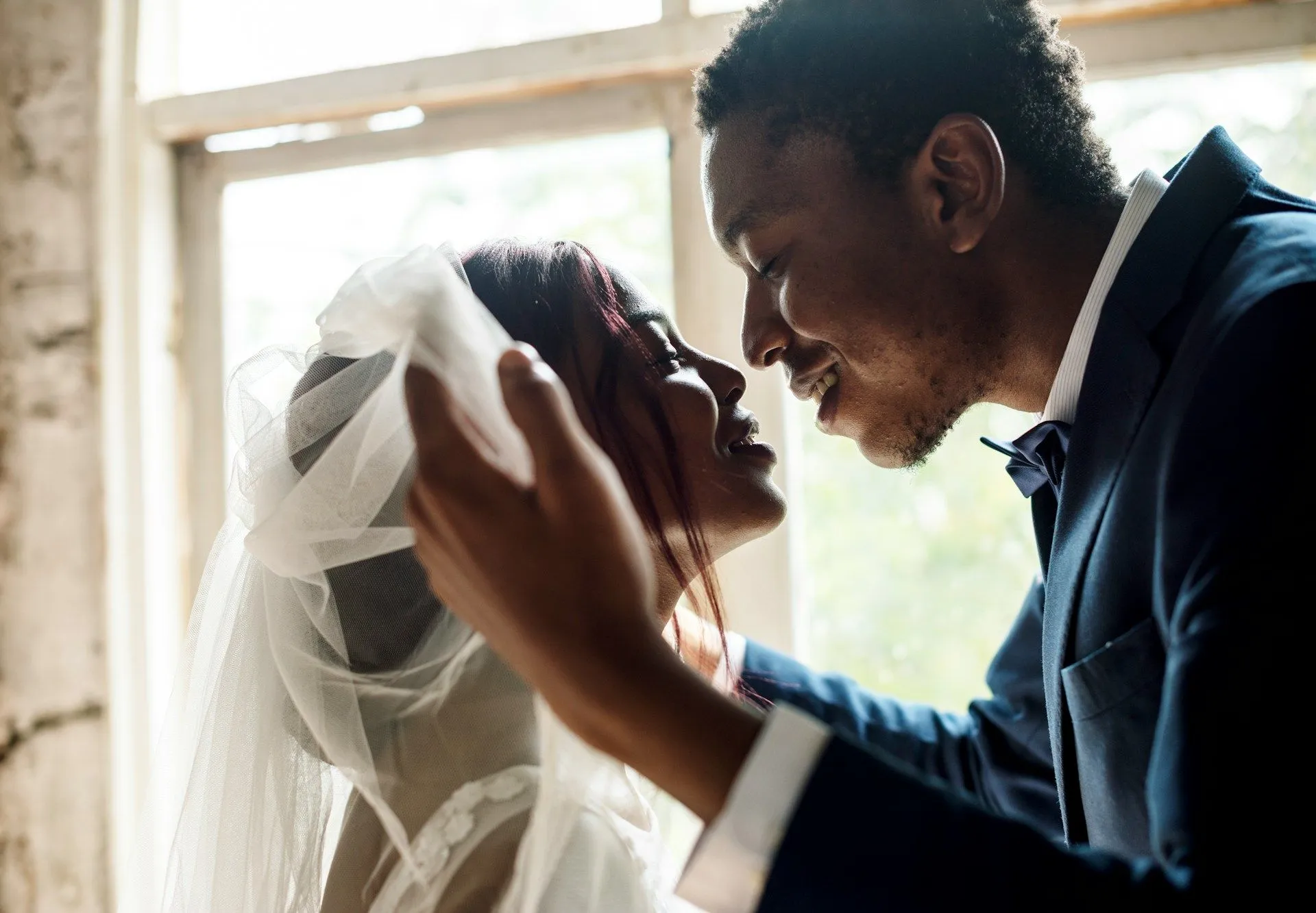 The bride and groom leaning in for a kiss by a sunlit window.