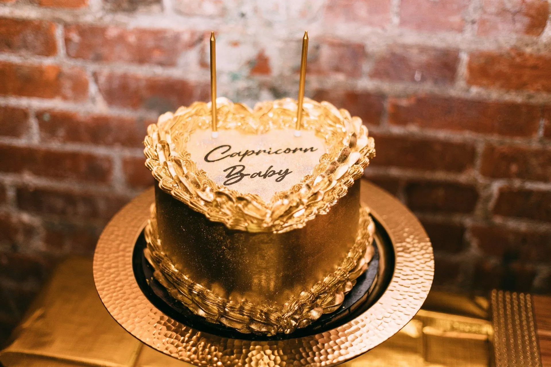 A gold heart-shaped cake with “Capricorn Baby” written on top and two candles.