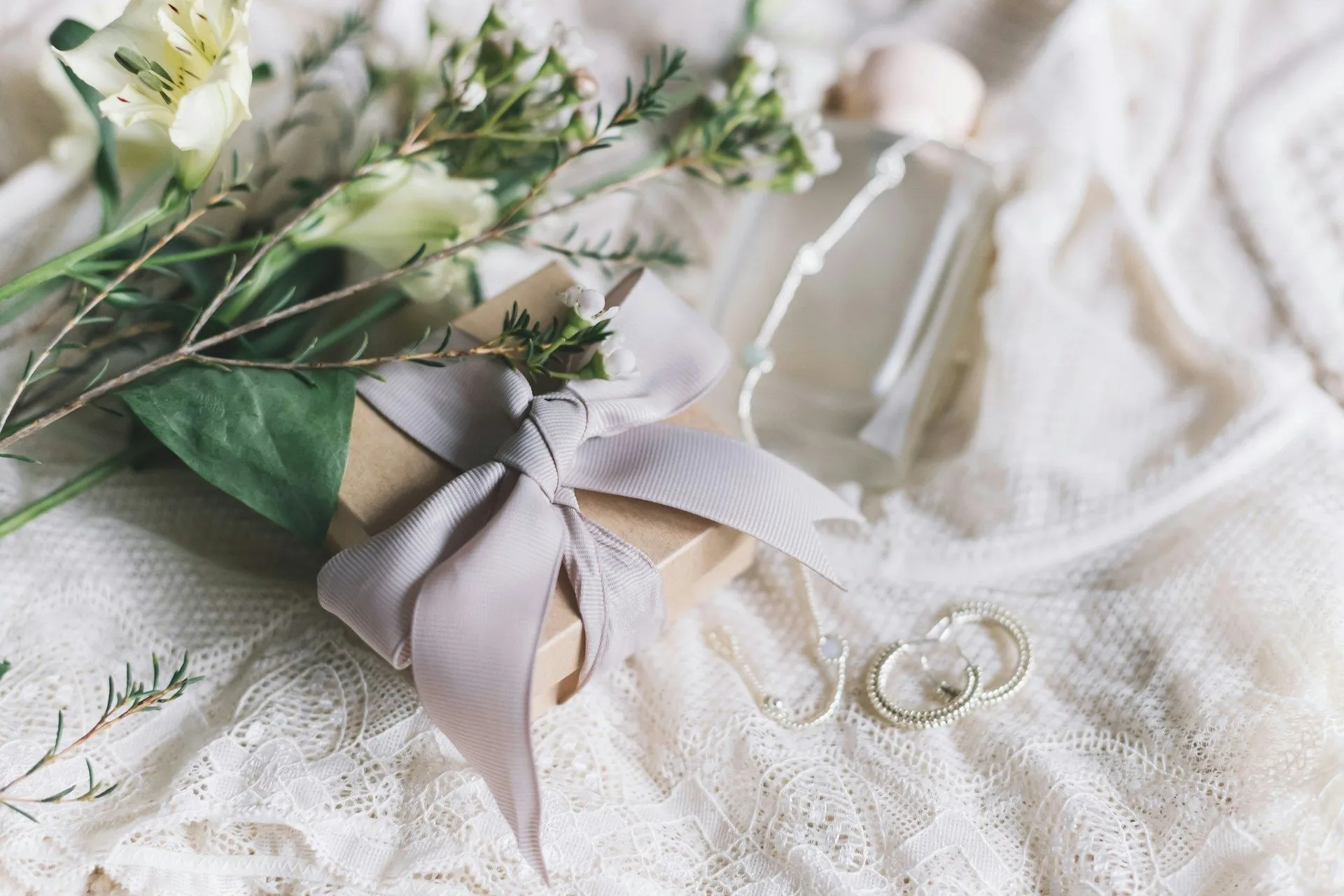 Gift box with a ribbon, flowers, perfume bottle, and jewelry on a lace fabric background.