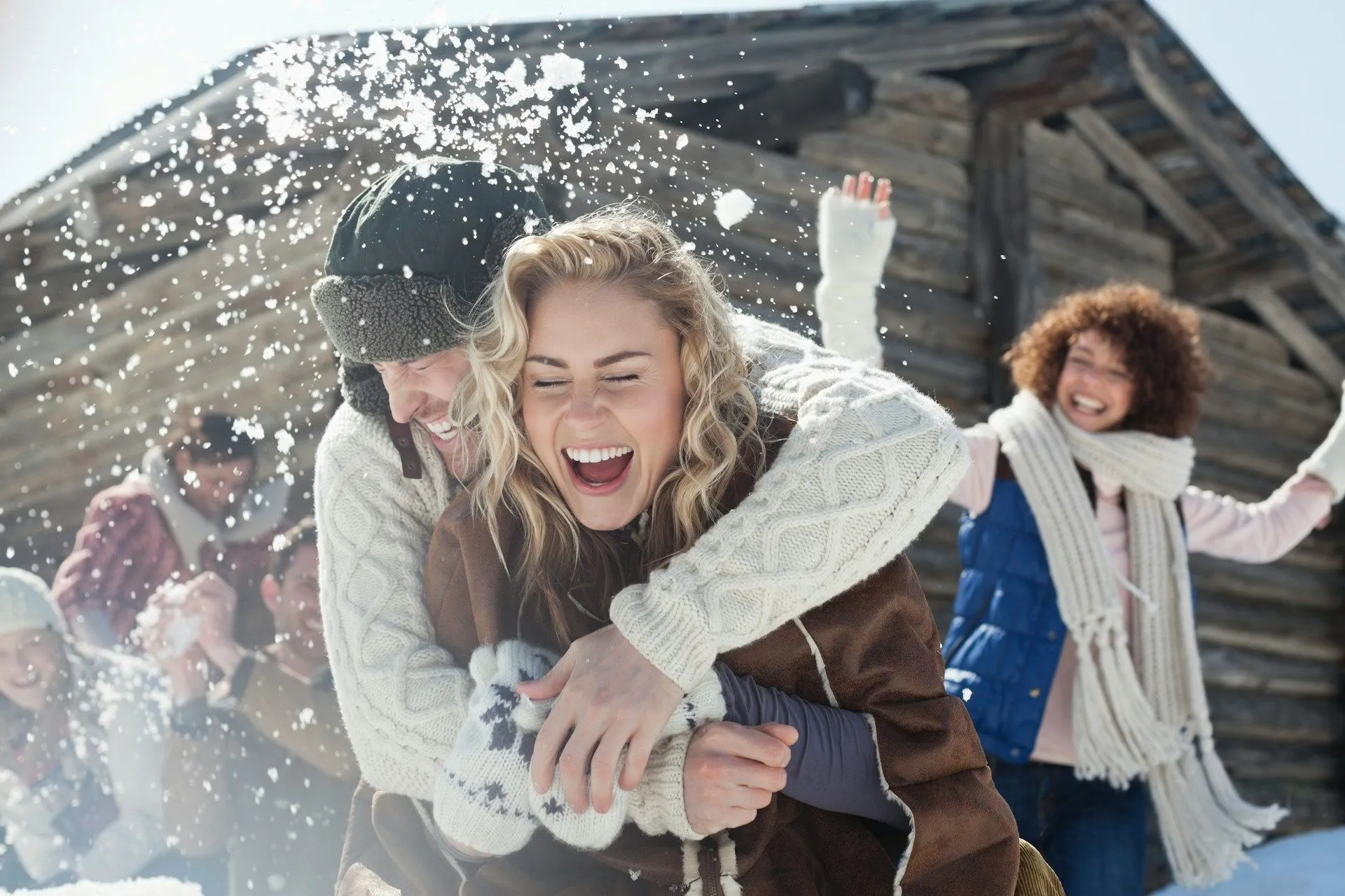 Friends playing in the snow and laughing outside a wooden cabin.