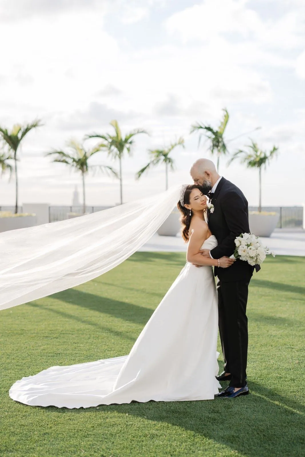 Outdoor wedding portrait of a bride and groom. Palm trees and a bright, sunny sky are in the background.