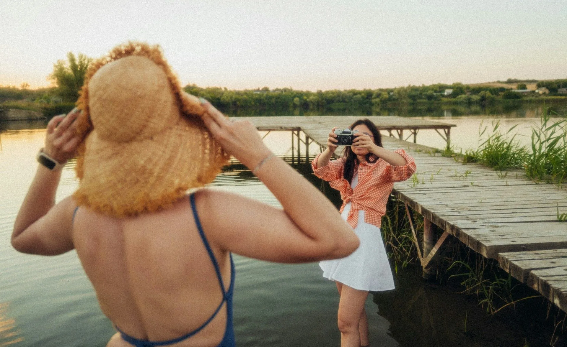 Two friends by a lake at sunset, one in a sun hat, the other taking a photo.