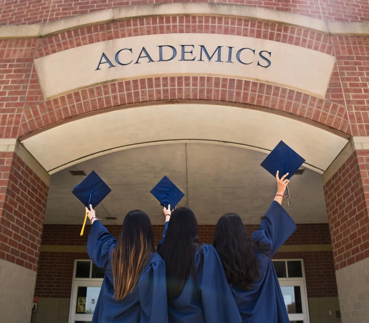 Three graduates in blue caps and gowns raise their mortarboards in front of a brick building entrance labeled “Academics.”