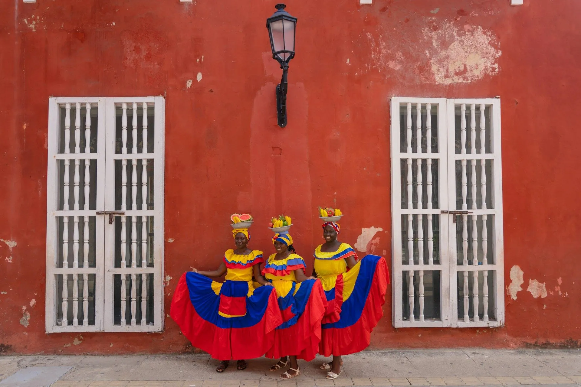 Three women in colorful traditional dresses pose against a red wall, holding fruit baskets on their heads.