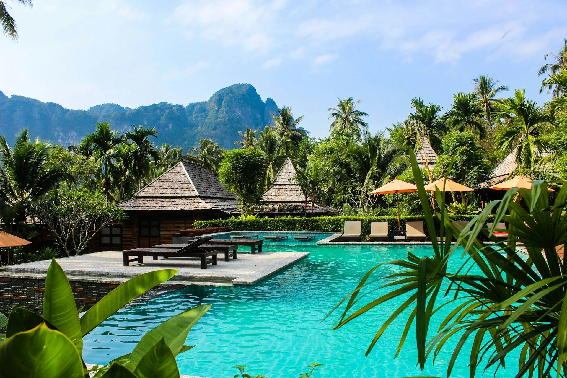 Resort pool surrounded by palm trees and tropical huts with mountains in the background.