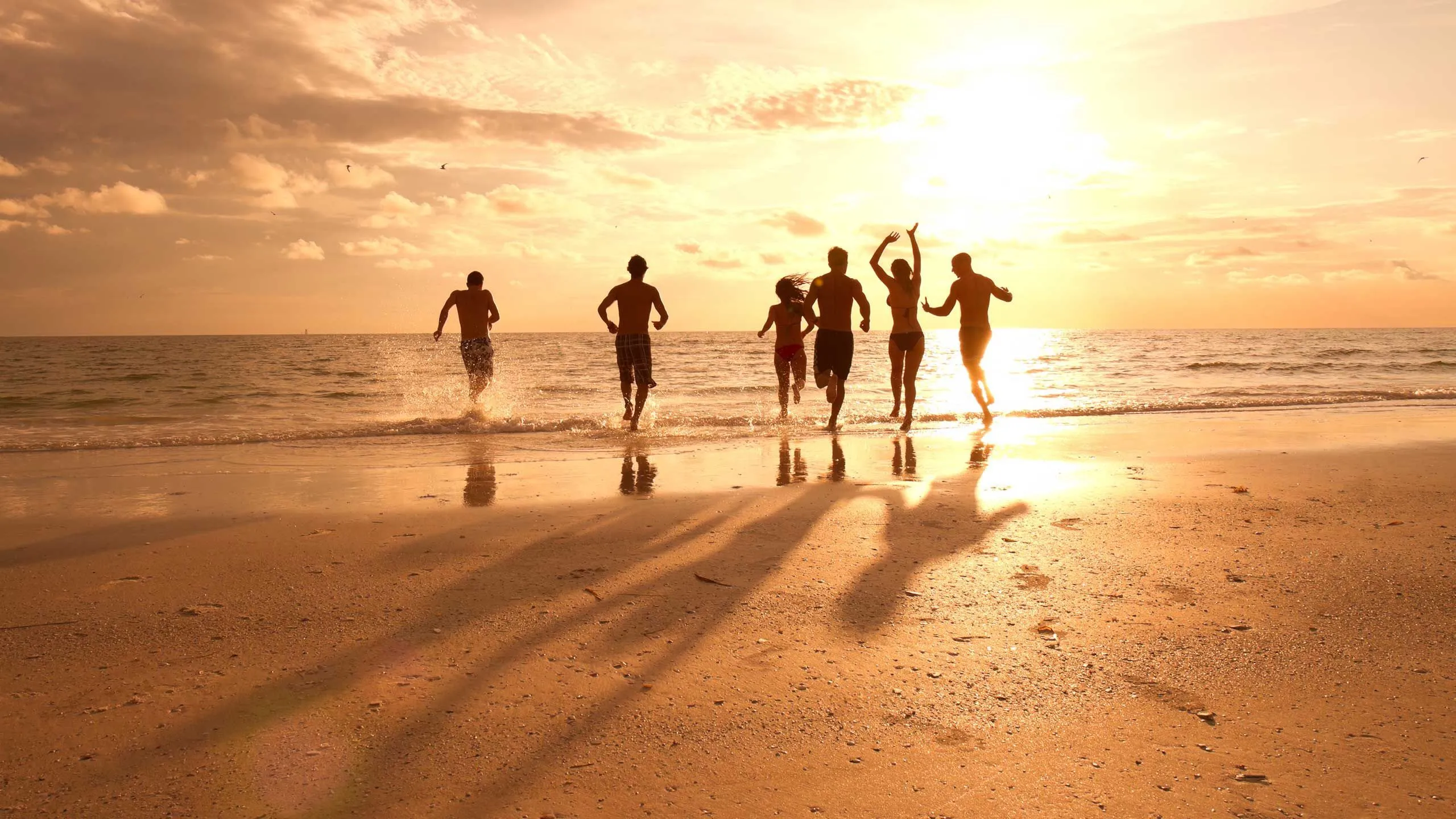 Friends running into the ocean together at sunset, casting long shadows on the beach