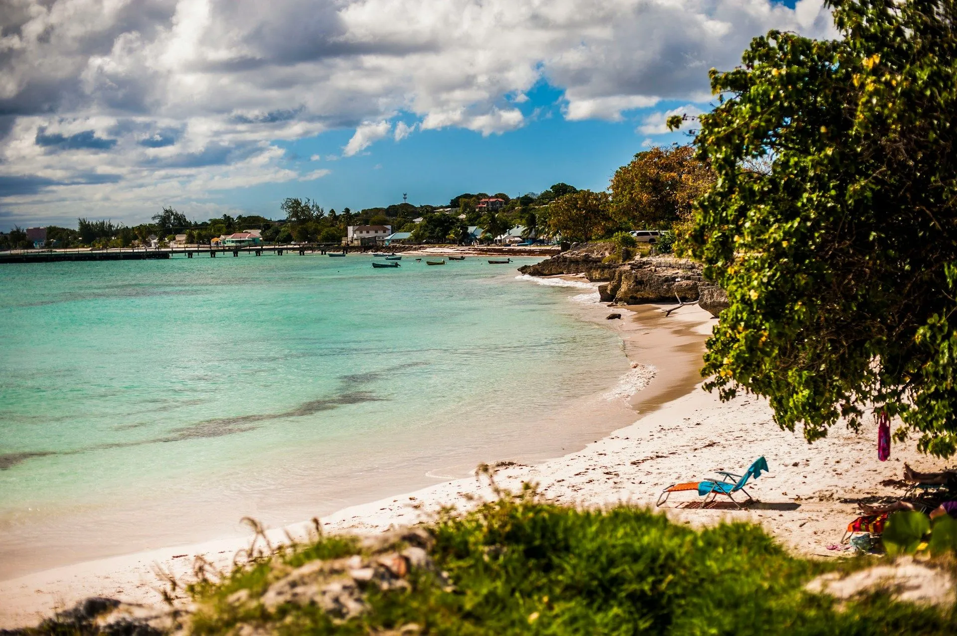 Calm turquoise water and pale sand along a quiet Barbados beach, with a wooden pier and lush green trees framing the shoreline.