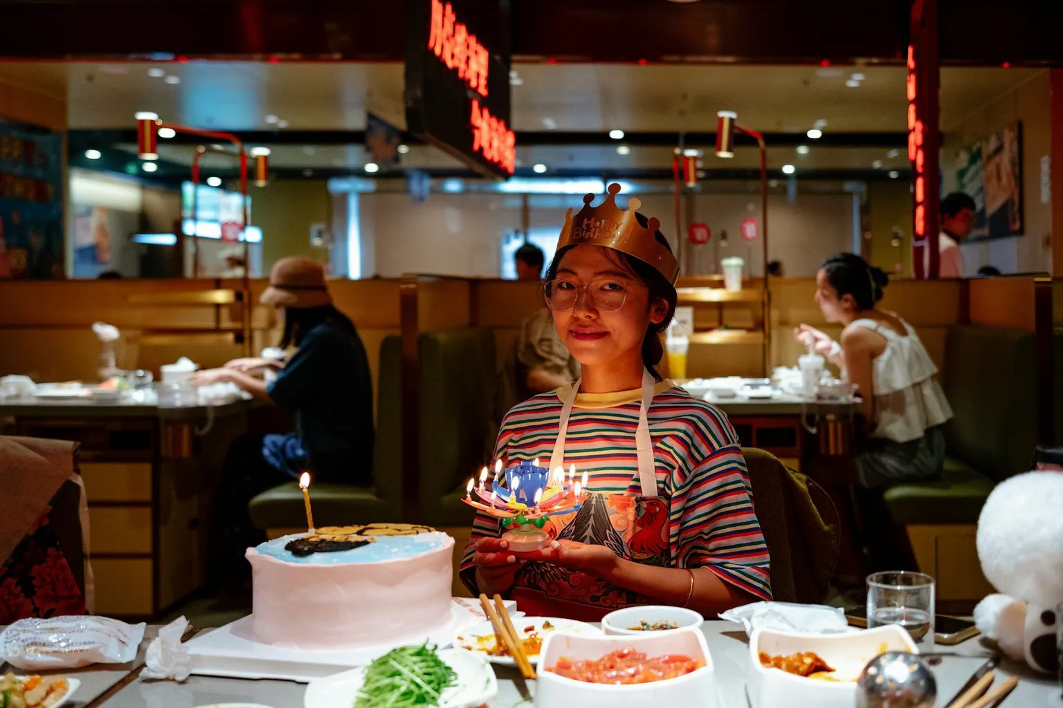 Woman smiling at a restaurant table holding a birthday cake with lit candles.